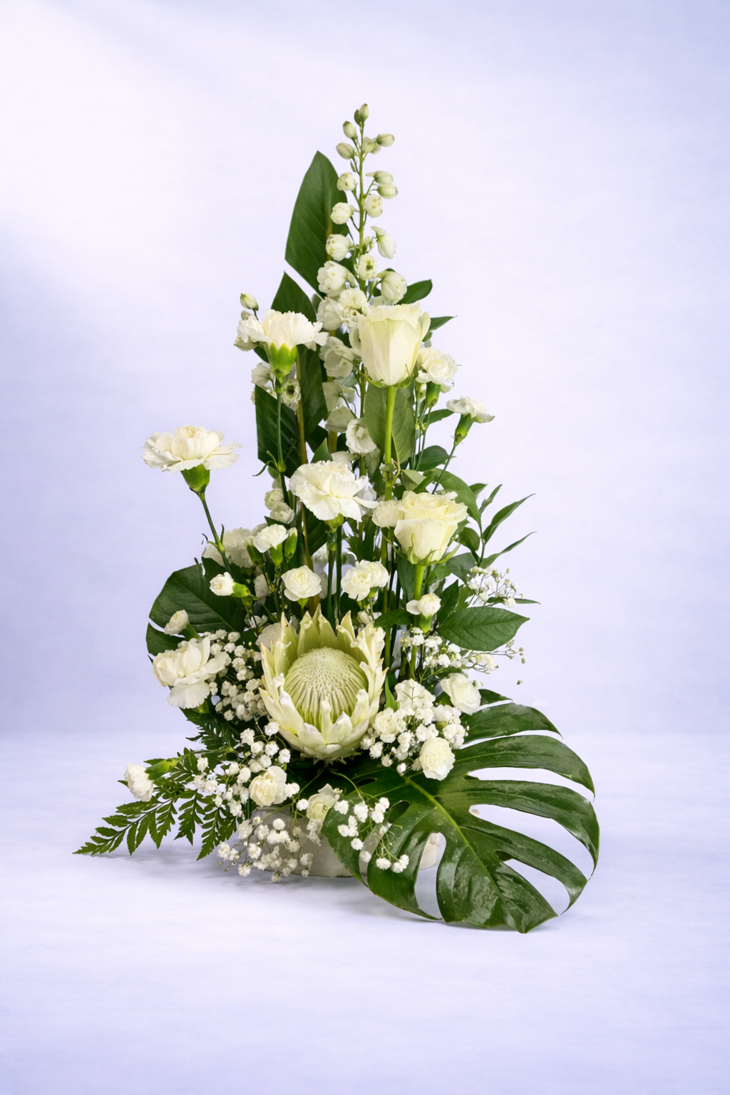 Elegant white floral arrangement featuring king protea, white roses, carnations and lush greenery on a soft neutral background.”