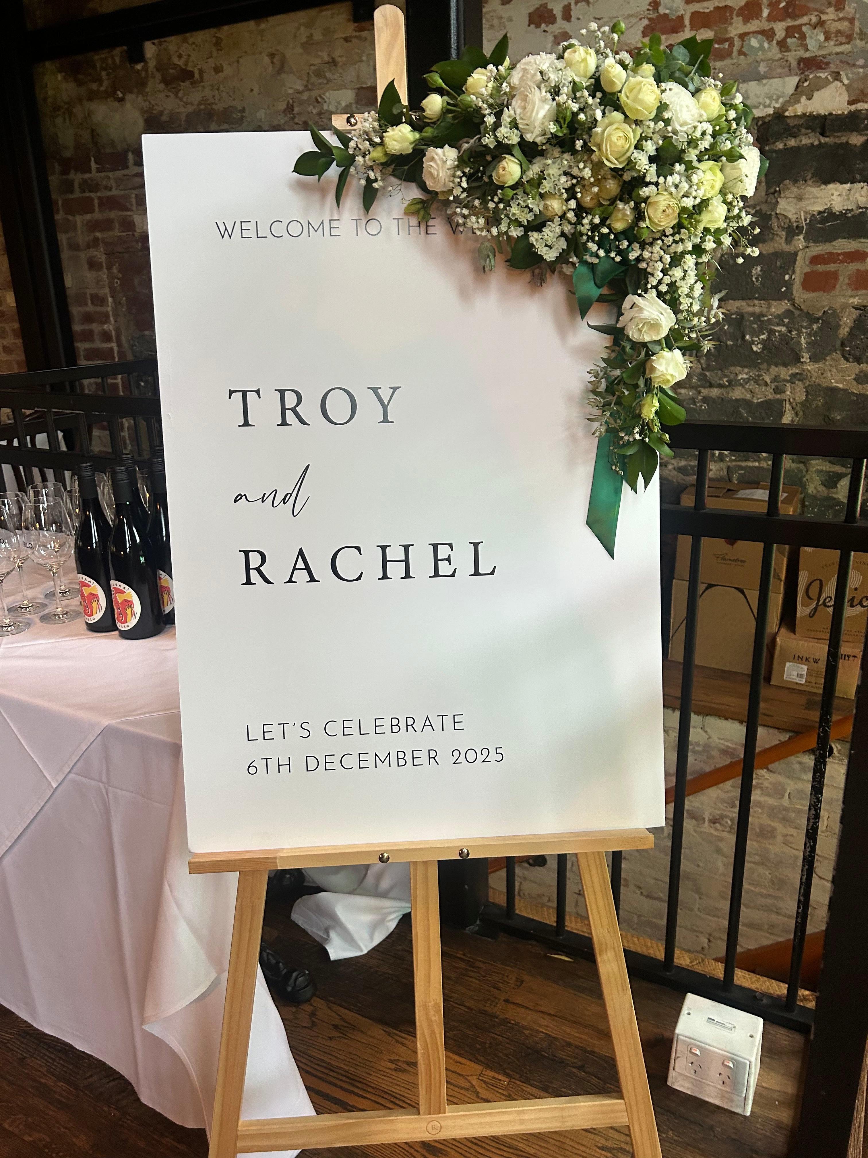 Wedding welcome sign with flowers on a stand against a brick wall.