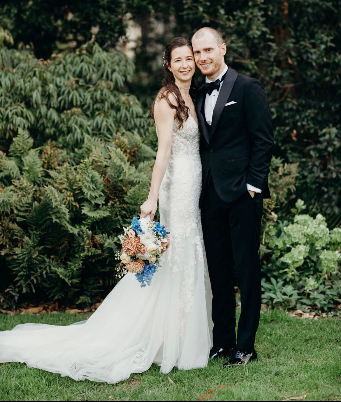Bride and groom standing in a garden, with the bride holding a bouquet featuring blue and peach wedding flowers by Emflowerment.