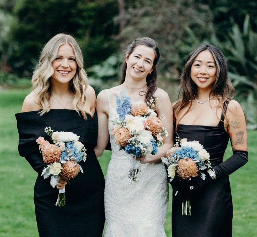 Three women, one in a wedding dress and two in black dresses, holding floral bouquets from Emflowerment florist.