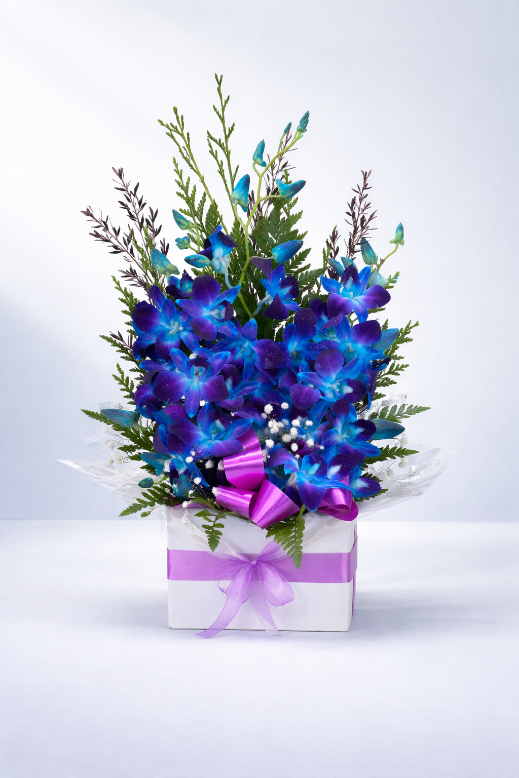 Vibrant blue Singapore orchids arranged in a white gift box with lush greenery, baby's breath, and a purple ribbon bow. A tall, elegant floral display perfect for birthdays, celebrations, or sympathy flowers.