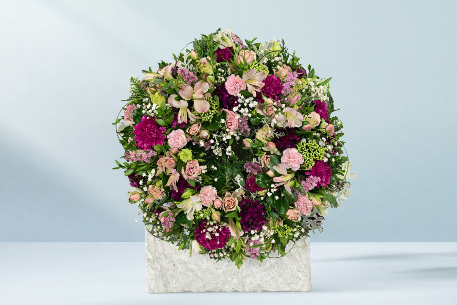 Round floral arrangement featuring pink roses, magenta carnations, lilies, baby’s breath and lush greenery in a white stone container.
